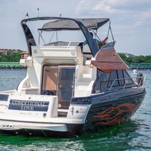 A motor yacht named "Conditional Love" on the water, featuring a canopy and visible cabins, with buildings in the background.