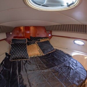 Interior of a boat cabin with a double bed, black bedding, patterned pillows, circular windows, and a round ceiling hatch.